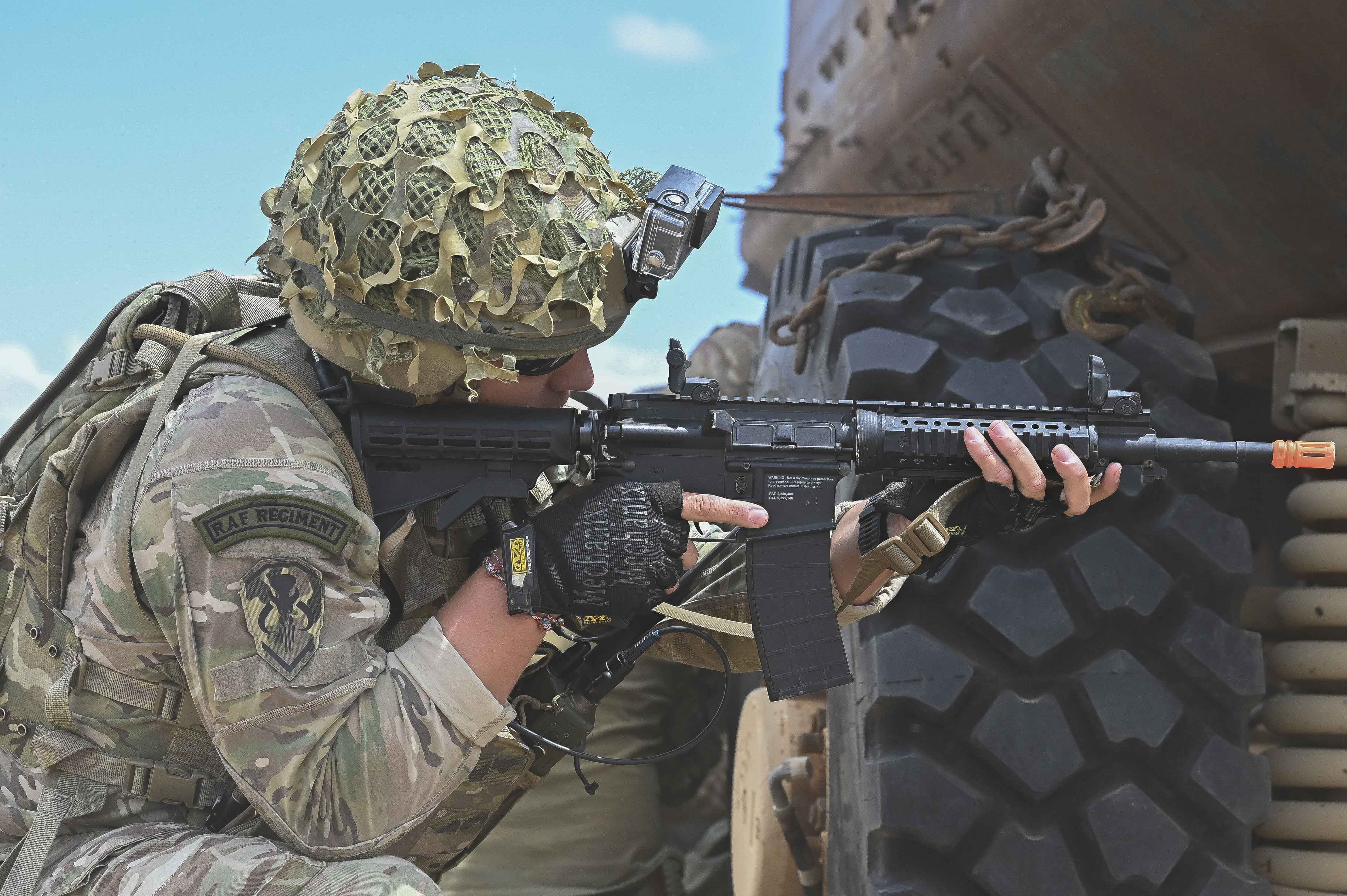 Image shows RAF Regiment with rifle, crouching by vehicle wheel.